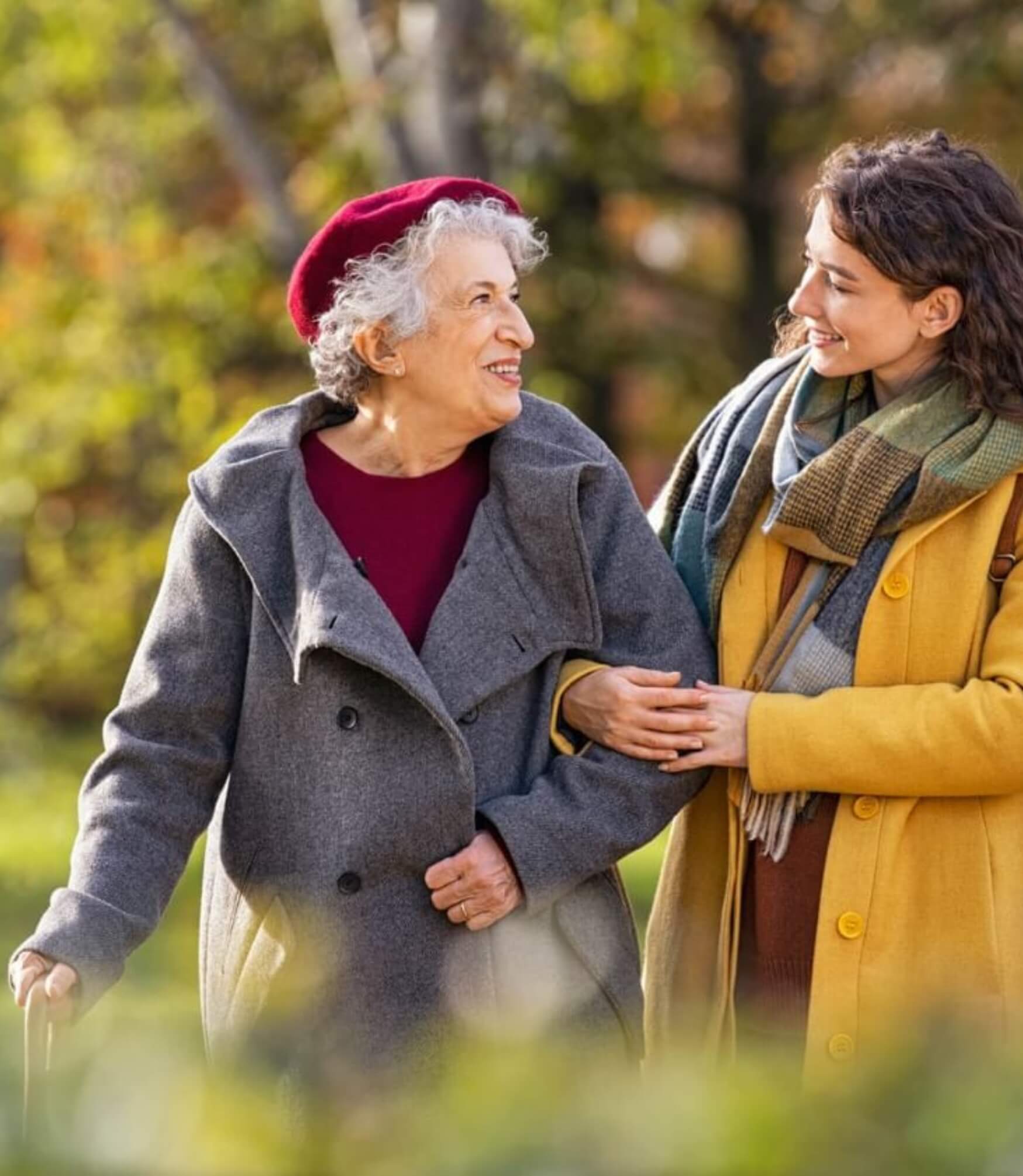 An older woman with a cane and a young woman smile at each other while walking outdoors on a sunny day. The elderly woman wears a grey coat and red beret, and the young woman wears a yellow coat.