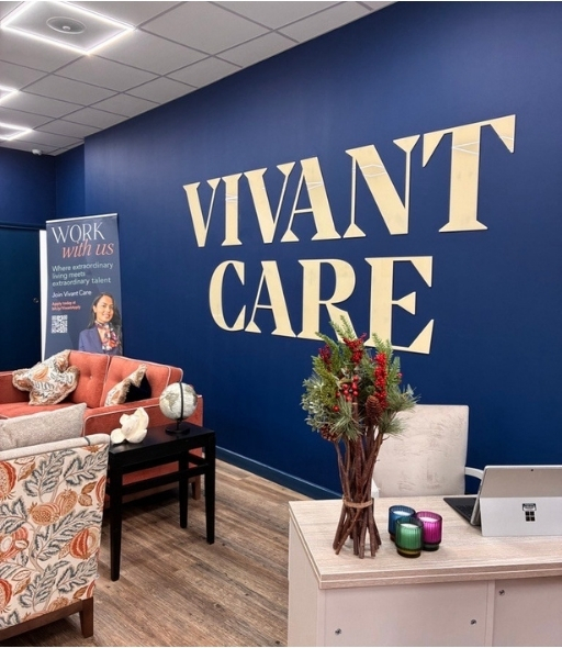 Modern Vivant Care Information Suite interior with deep blue wall featuring large gold 'VIVANT CARE' lettering. The space includes a desk with colorful candles and a festive floral arrangement, along with comfortable seating in patterned and coral tone.
