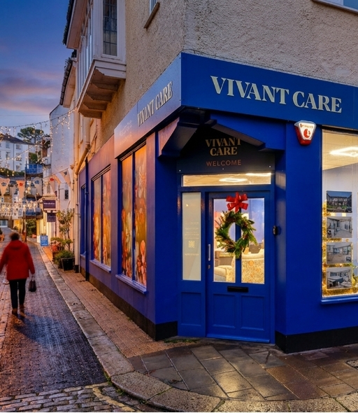 Street view of a corner building with bright blue exterior signage reading “VIVANT CARE.” The entrance door is decorated with a festive wreath and red bow, and large windows display vibrant images. A person is walking along the cobblestone street carrying a handbag, with warm evening lighting and decorative string lights visible in the background.