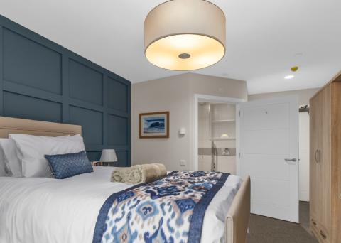 Bright, modern care home bedroom featuring a large bed with white linens, blue accent pillows and a patterned throw, a dark blue feature wall, soft overhead lighting, and an open door leading to an en-suite bathroom